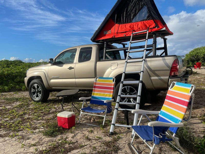 A tan pickup truck with a red rooftop tent and ladder, set up as an rv rental for camping. Beach chairs and cooler on sandy ground. — photo 18