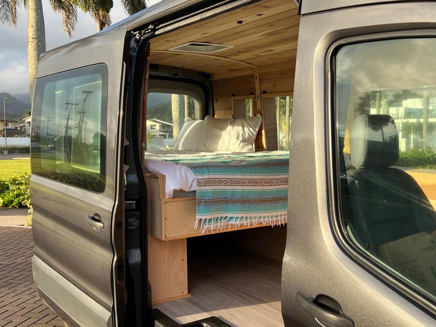 RV rental: A custom grey camper van with its side door open, revealing a cozy bed and warm wooden interior, ready for travel.