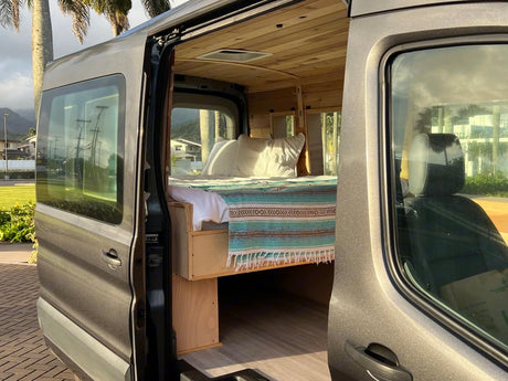 RV rental: A custom grey camper van with its side door open, revealing a cozy bed and warm wooden interior, ready for travel.