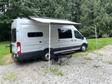 Light gray camper van with awning & folding chair, ready for your rv rental adventure in nature.