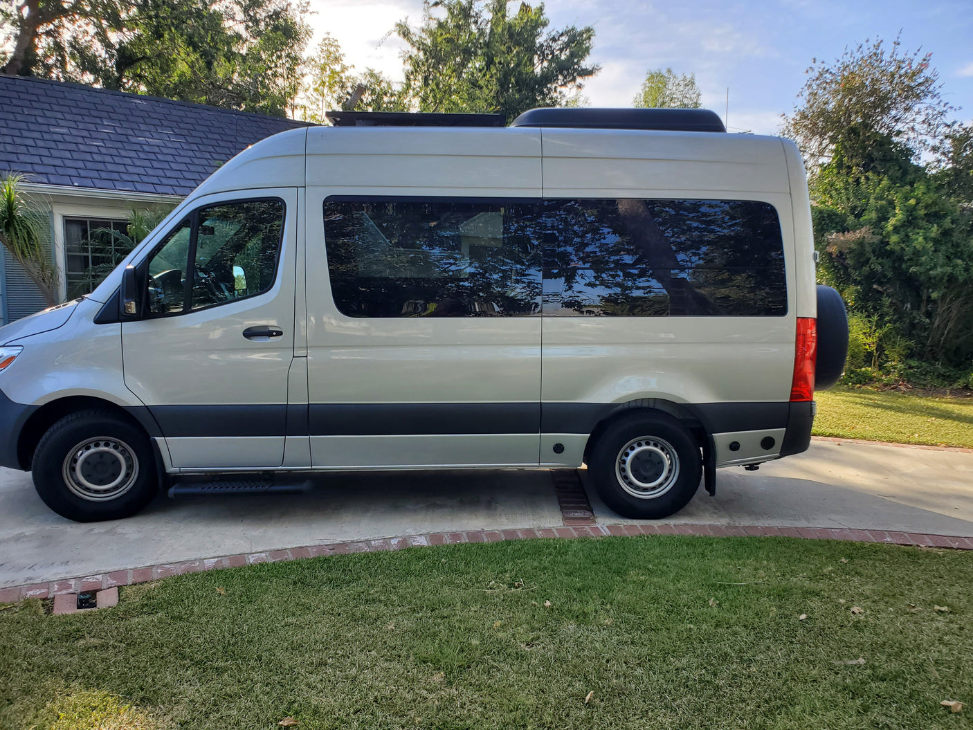 Side view of a light-colored camper van for RV rental, parked on a driveway beside a house and green lawn.