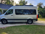 Side view of a light-colored camper van for RV rental, parked on a driveway beside a house and green lawn.