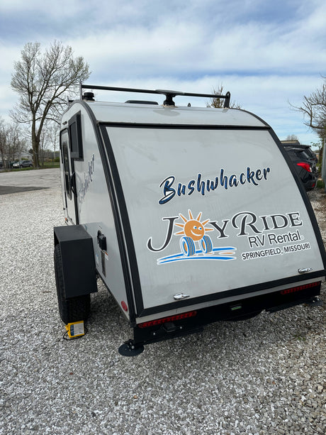 Rear view of a light gray Bushwhacker RV rental, branded JoyRide RV Rental, parked on gravel.