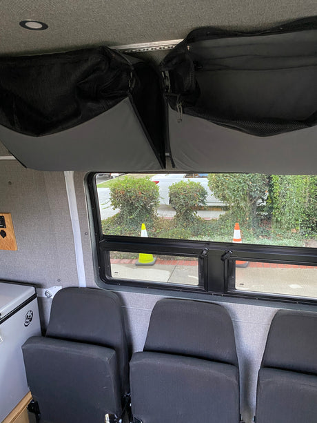 RV rental: Overhead mesh storage, window to outdoor traffic cones/bushes, passenger seats, and cooler visible.