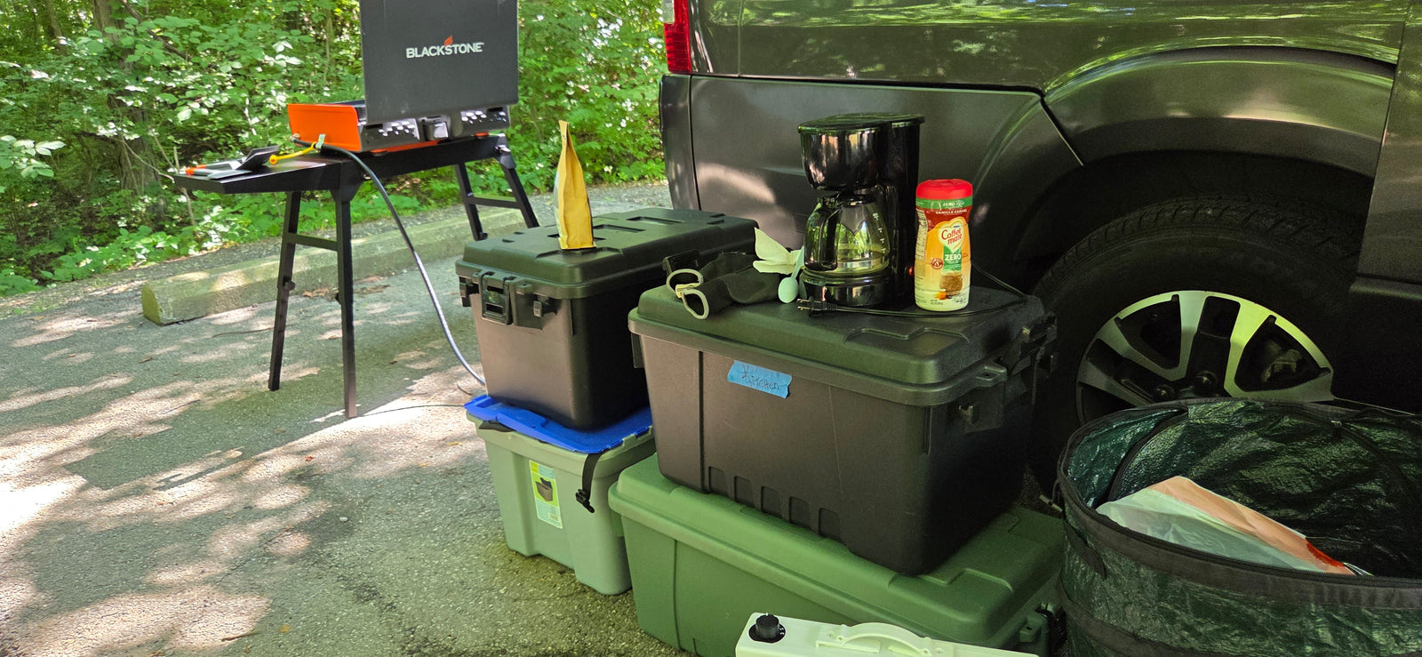 Outdoor kitchen setup featuring a Blackstone griddle, coffee maker, and storage bins next to an RV rental vehicle for camping convenience.