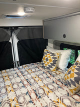 Cozy RV rental interior: bed with patterned comforter & pillows, overhead light, vent, and screened rear doors.