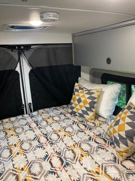 Cozy RV rental interior: bed with patterned comforter & pillows, overhead light, vent, and screened rear doors.