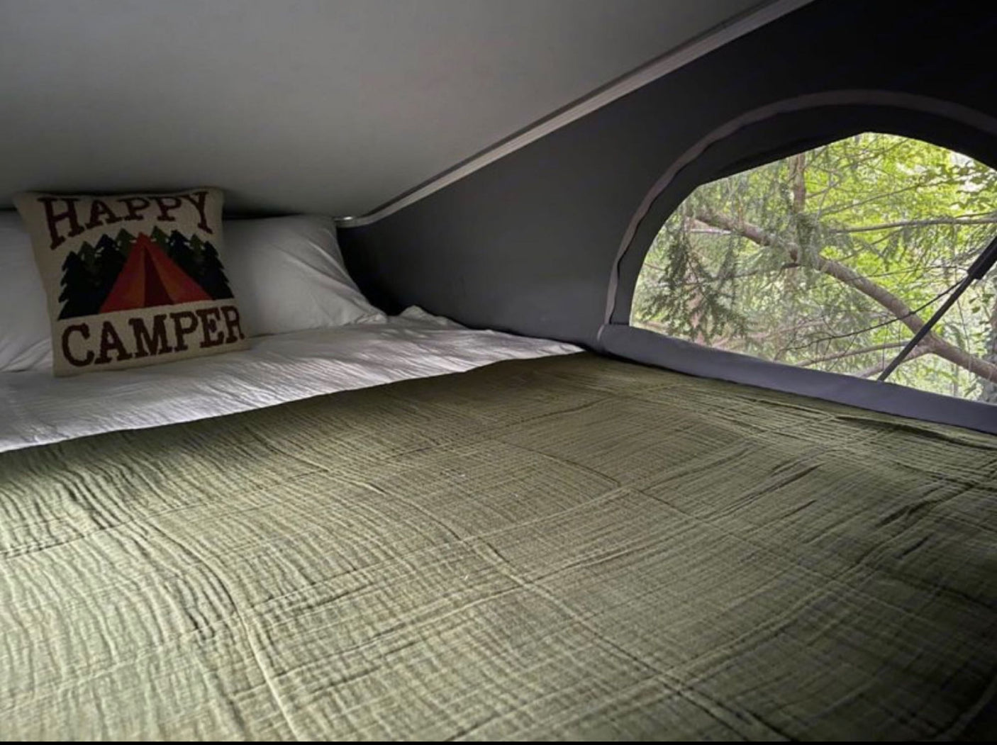 Cozy RV rental bed with a "Happy Camper" pillow and a scenic view of lush green trees from the large window.