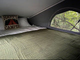 Cozy RV rental bed with a "Happy Camper" pillow and a scenic view of lush green trees from the large window.