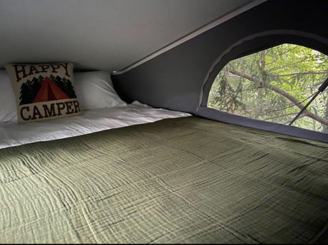 Cozy RV rental bed with a "Happy Camper" pillow and a scenic view of lush green trees from the large window.
