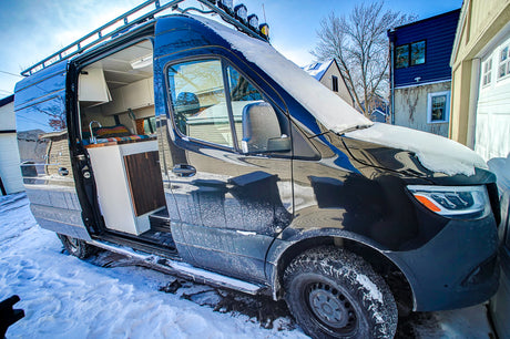 Black RV rental van with side door open, revealing its interior, parked in a snowy winter setting.