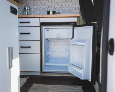 RV rental kitchen showcasing a compact, open refrigerator, white cabinets, wood counter, and tile backsplash.