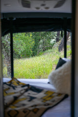 A view from inside an RV rental bed reveals vibrant green nature: trees, grass, and rocks outside the window.