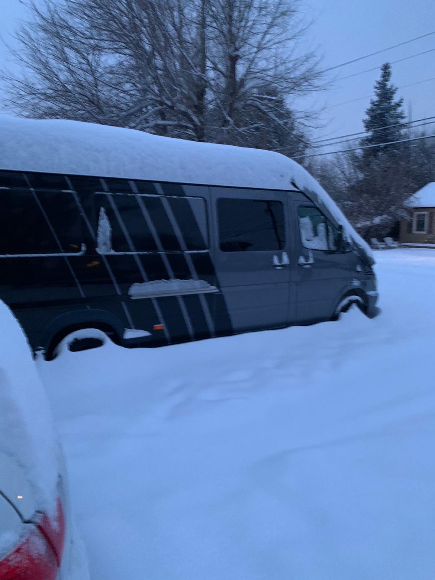 A gray RV rental van is almost completely buried in deep snow, highlighting extreme winter conditions. Bare trees surround it.