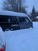 A gray RV rental van is almost completely buried in deep snow, highlighting extreme winter conditions. Bare trees surround it.