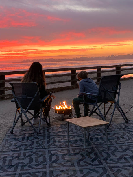 Amazing RV rental adventure ends with a fiery beach campfire. Two people enjoy the vivid red and orange sunset over the ocean.