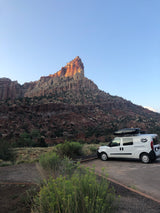 An rv rental van parked at the base of a towering, sun-kissed rock formation in a desert landscape.