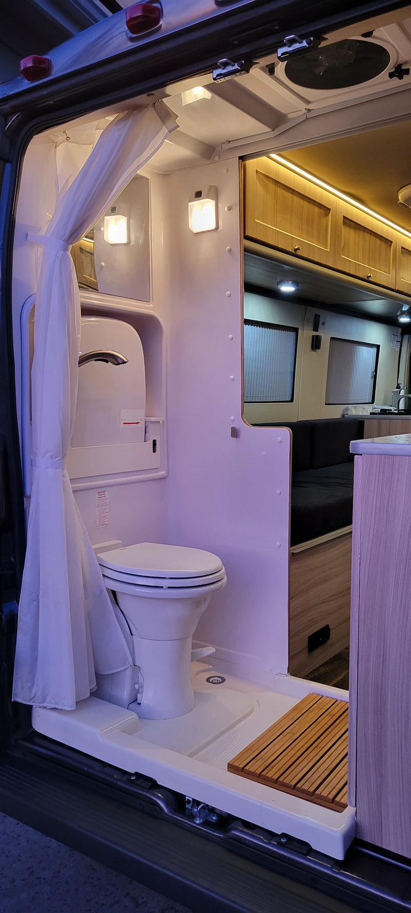 RV rental interior: A compact, illuminated camper van bathroom with toilet, shower, and adjacent kitchen/living area. — photo 8