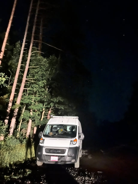 White RV rental van parked on a forest road at night. Tall trees and a person's shadow are visible under a starry sky.