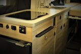 Bright RV rental kitchen: a sleek stovetop, sink, Novacool fridge, and useful power ports on light wood cabinets.