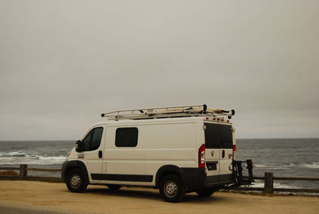 White RV rental camper van parked on a dirt road next to the ocean, with waves crashing. Ideal for coastal travel.