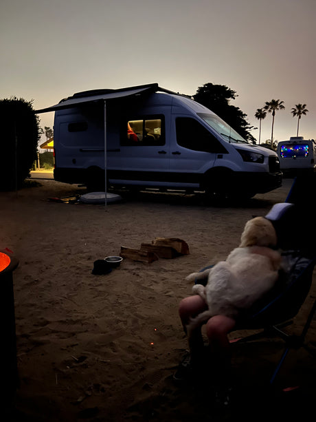 Relaxing at an RV rental campsite at dusk. A person with a dog on lap, fire pit, and camper van with awning.