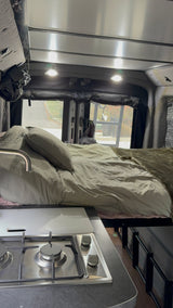 Inside an RV rental, a bed with light bedding is next to a compact kitchen with a two-burner stove and sink faucet.