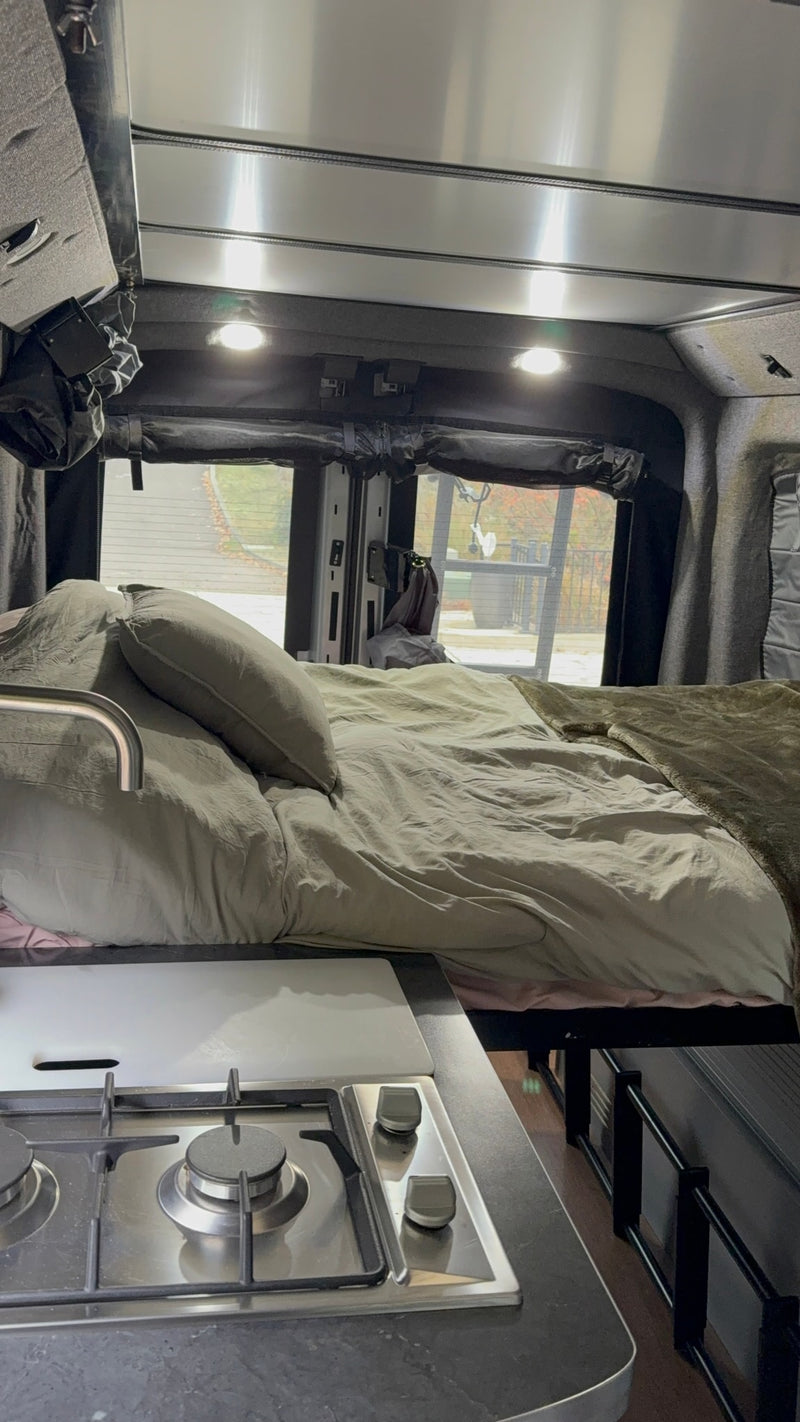 Inside an RV rental, a bed with light bedding is next to a compact kitchen with a two-burner stove and sink faucet. — photo 11