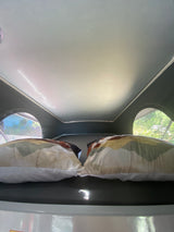 RV rental interior: A bed with two pillows and rounded windows showing outside trees.
