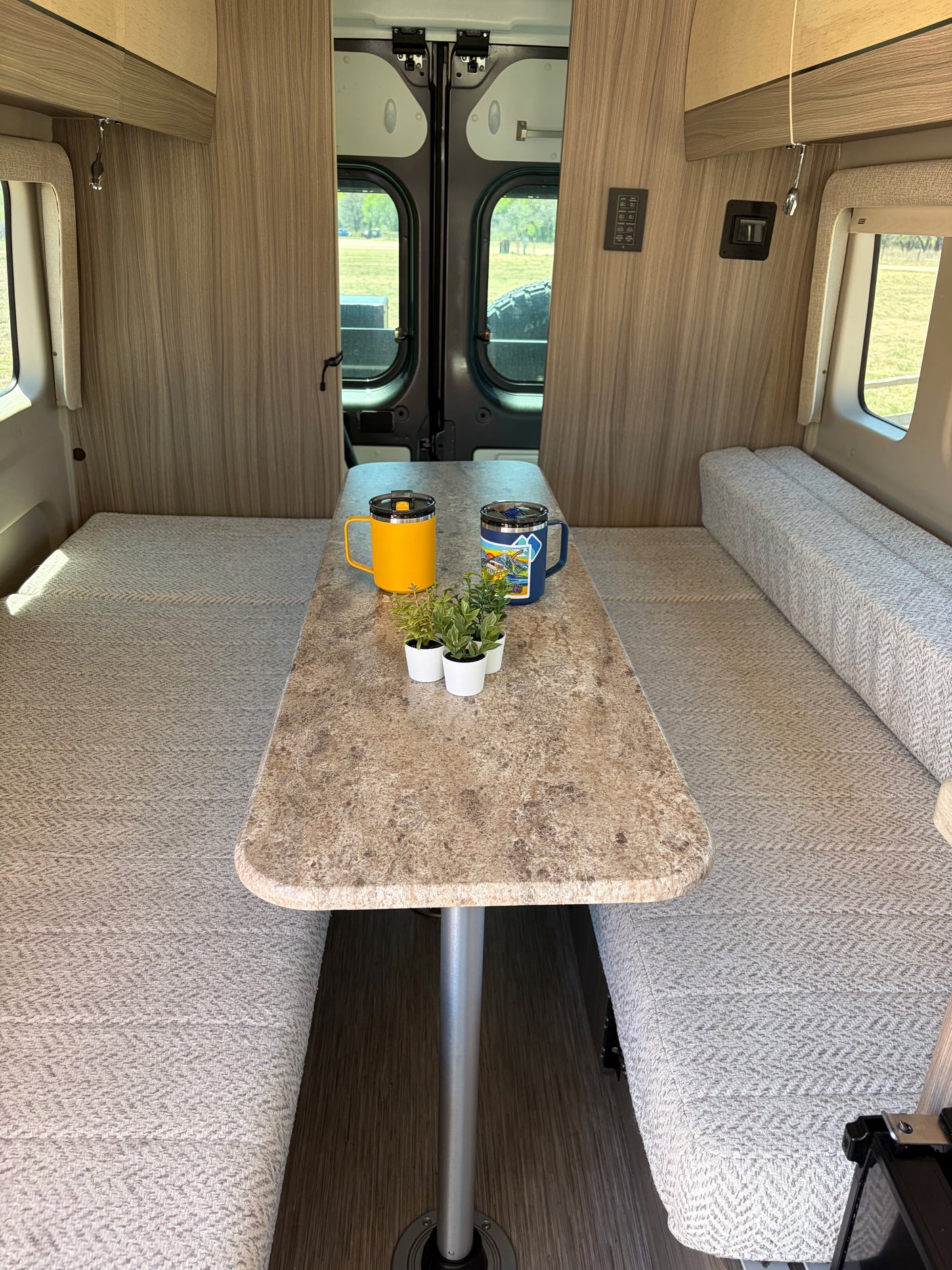 Cozy RV rental interior features a dining area with wood-paneled walls, comfy benches, a table, mugs, and plants.
