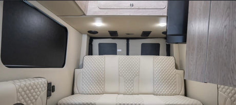 Modern rv rental interior: light-colored quilted seating, overhead cabinets, and dark window blinds. Ready for travel.