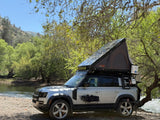 Custom Land Rover Defender w/ rooftop tent & gear by river. RV rental for overlanding adventures.