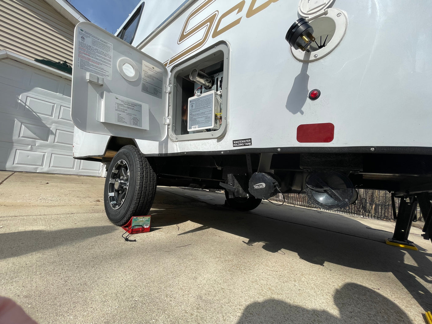 Side view of an RV rental with service door open, revealing utilities, wastewater tank connection, and wheel chocks during maintenance.