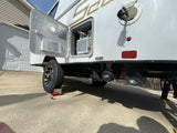 Side view of an RV rental with service door open, revealing utilities, wastewater tank connection, and wheel chocks during maintenance.