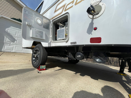 Side view of an RV rental with service door open, revealing utilities, wastewater tank connection, and wheel chocks during maintenance.