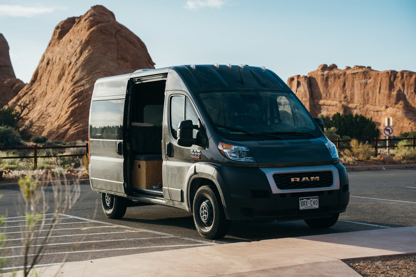 Grey Ram ProMaster RV rental van with open side door, parked in a desert landscape with large red rock formations.
