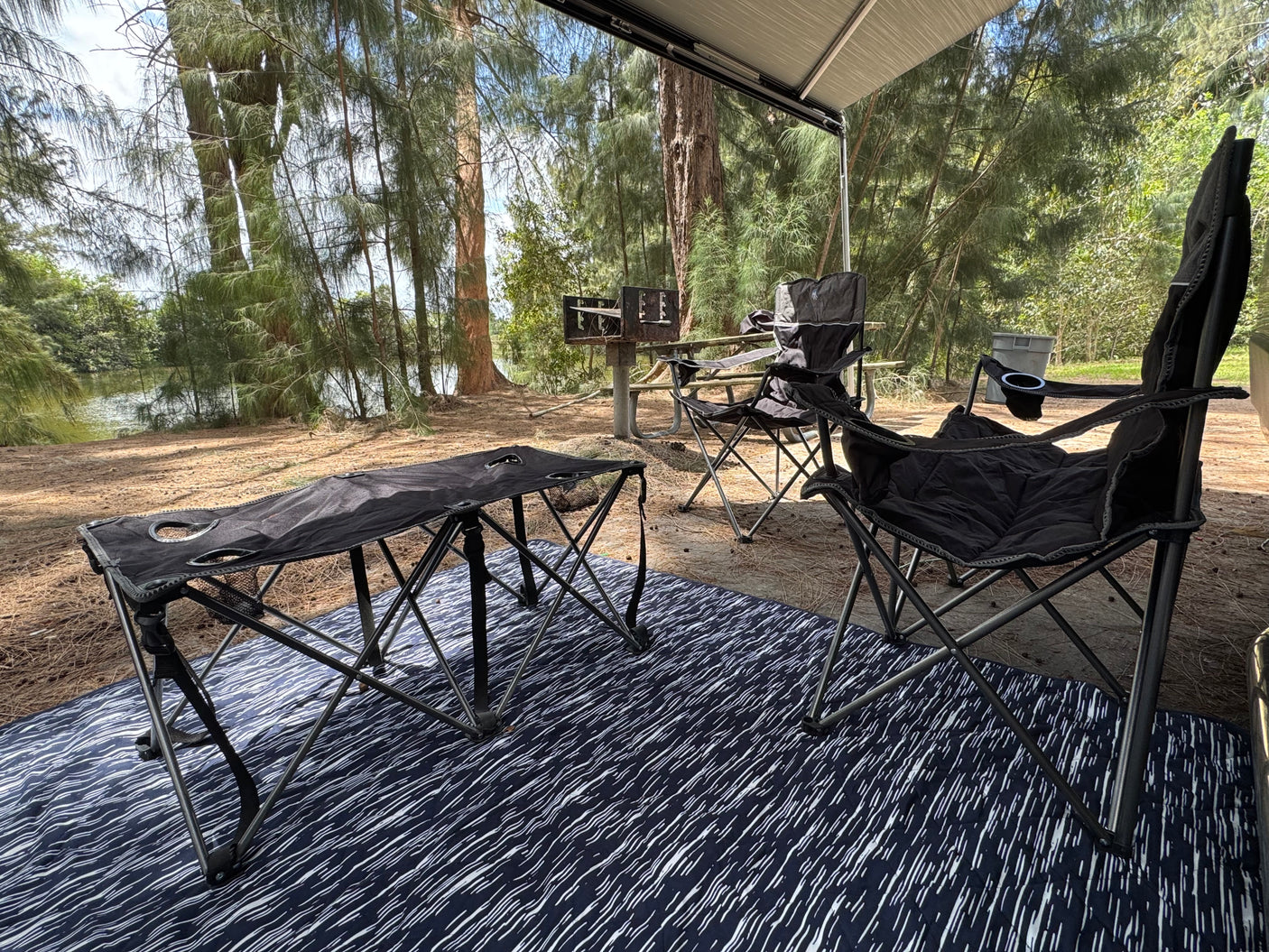 Peaceful RV rental campsite with awning, comfy chairs, and table on a mat, overlooking a BBQ grill and serene waters.