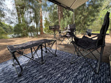 Peaceful RV rental campsite with awning, comfy chairs, and table on a mat, overlooking a BBQ grill and serene waters.
