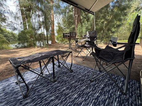 Peaceful RV rental campsite with awning, comfy chairs, and table on a mat, overlooking a BBQ grill and serene waters.