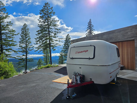 A white Scamp travel trailer, ideal for RV rental, overlooks a misty mountain valley from a modern driveway under a bright sky.