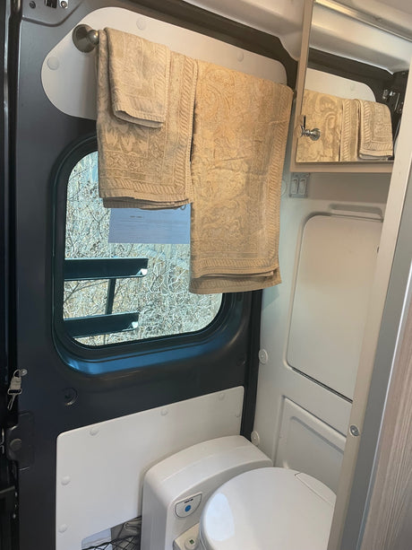Compact RV rental wet bath interior, featuring a toilet, hanging towels, and a window overlooking trees.