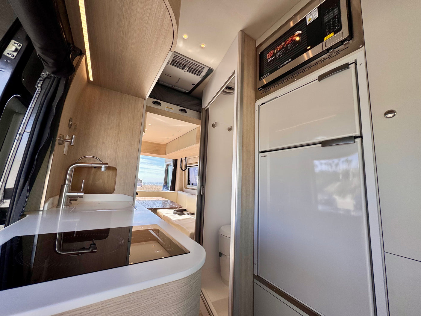 Modern RV rental interior with a compact kitchen (sink, cooktop), built-in fridge, microwave, and ocean view.