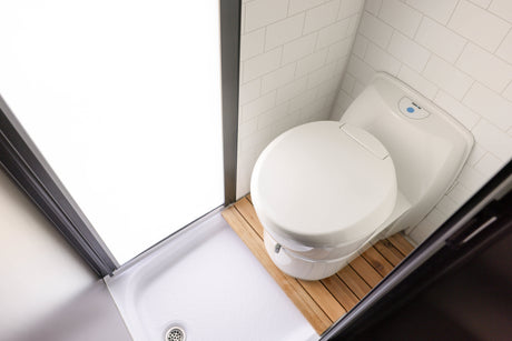 Modern RV rental wet bath with a compact white toilet on a wood mat, white tiled walls, and shower pan. Space-saving for travel.