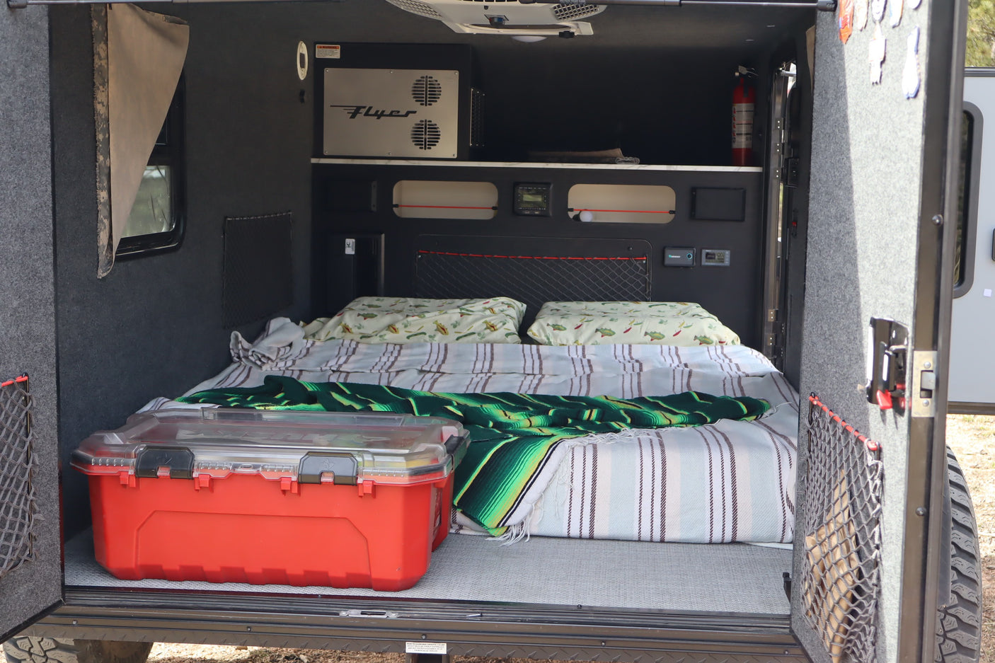 Cozy rv rental camper interior with a bed, pillows, blankets, and a red storage box. Controls and a fire extinguisher are visible.