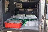 Cozy rv rental camper interior with a bed, pillows, blankets, and a red storage box. Controls and a fire extinguisher are visible.