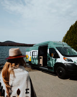 A person by a lake admires a custom green camper van (RV rental) with mountain graphics, ready for outdoor adventure.
