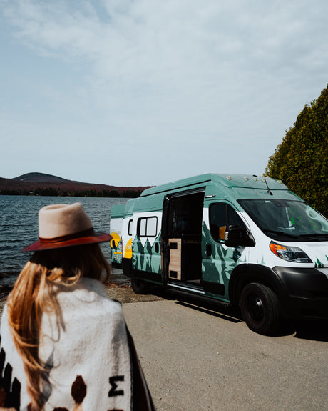 A person by a lake admires a custom green camper van (RV rental) with mountain graphics, ready for outdoor adventure.