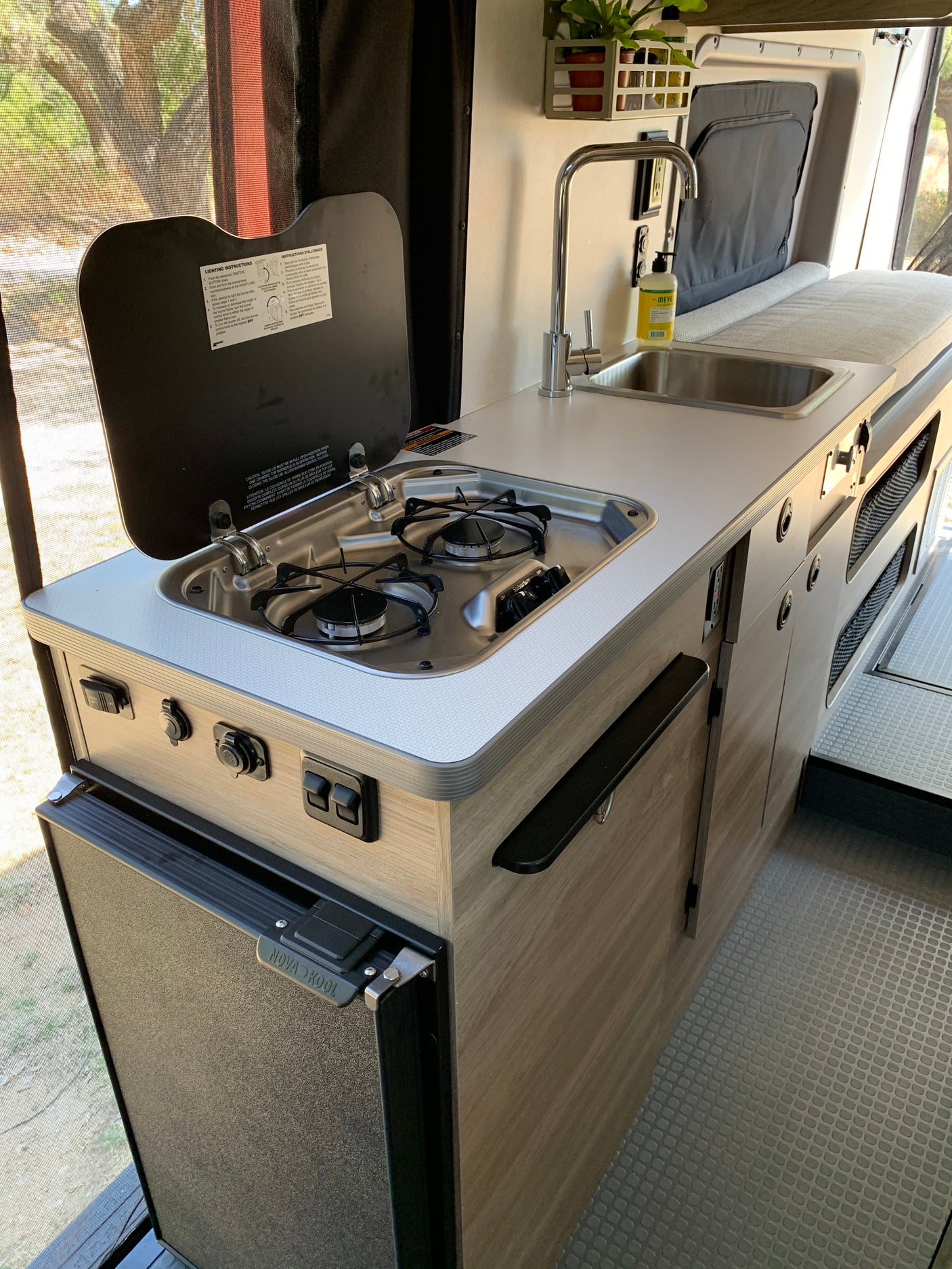 Inside an RV rental: a compact kitchen with a two-burner stove, sink, and Nova Kool refrigerator, ready for your adventure.