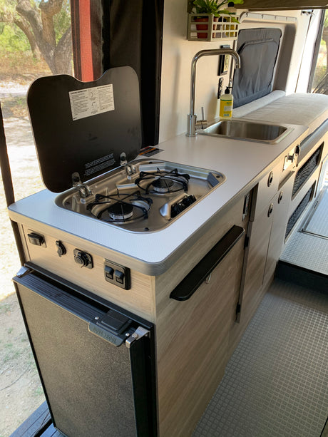 Inside an RV rental: a compact kitchen with a two-burner stove, sink, and Nova Kool refrigerator, ready for your adventure.
