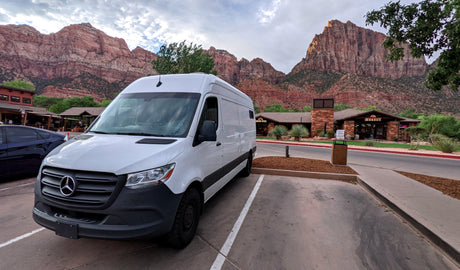 White Mercedes Sprinter RV rental parked in Zion National Park, with towering red mountains and village buildings in the background.